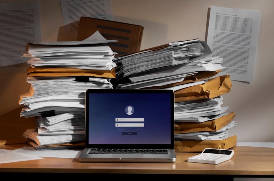 Uma mesa desorganizada com pilhas de documentos e um laptop com tela de login, simbolizando a necessidade de digitalização e otimização da gestão documental no RH.